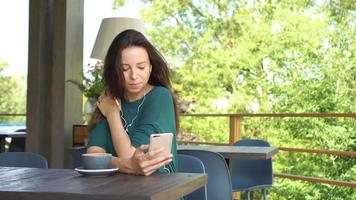 Frau, die App auf dem Smartphone im Café verwendet, trinkt Kaffee, lächelt und schreibt auf dem Handy eine SMS. schöne junge weibliche Fachkraft mit Handy. video