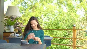 jovem mulher com smartphone enquanto está sentado sozinho na cafeteria durante o dia quente. mulher atraente com sorriso fofo escrevendo mensagem com telefone celular enquanto descansa no café video