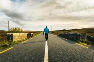 Back view male cyclist in blue jacket walk on road by touring bicycle travel outdoors in Ireland. Purpose and travel adventure concept. Wild atlantic way road trip photo