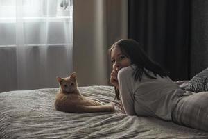 una hermosa joven en pijama yace en una cama en casa, junto a su gato británico de durazno foto