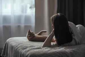 A sad young woman with seasonal affective disorder lies alone on the bed and looks out the window, next to a domestic cat. Concept of winter depression due to lack of sunlight, selective focus photo