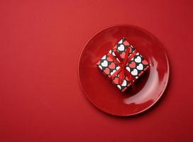 square cardboard box tied with a silk red ribbon lies in a round ceramic red plate, top view photo