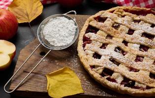 baked round traditional apple pie on a brown wooden board and fresh red apples. Next to it is an iron strainer with white powdered sugar photo