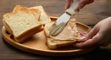 female hand spreads peanut butter on a square slice of white wheat flour photo