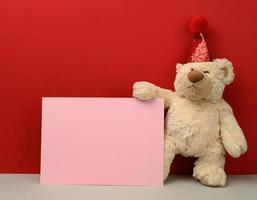 teddy bear in a red festive hat holds a pink sheet of paper on a red background photo