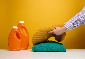 ropa doblada lavada y botellas grandes de plástico naranja con detergente líquido sobre una mesa blanca, fondo amarillo. tarea de rutina foto