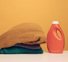 pila de ropa doblada lavada y botella grande de plástico naranja con soporte de detergente líquido sobre una mesa blanca, fondo amarillo. tarea de rutina foto