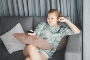 Asian housewife sit in sofa and holding remote control watching television with relaxing action. Hobby and free time life style concept. photo