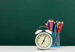 round alarm clock and multicolored pencils on the background of an empty green chalk board. Back to school photo