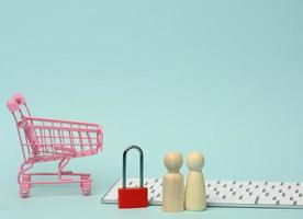 a red metal padlock stands on the keyboard and behind a miniature shopping cart, blue background photo