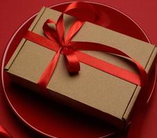 rectangular brown cardboard box tied with a silk red ribbon lies in a round ceramic red plate, top view photo