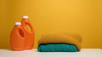 stack of washed folded clothes and plastic orange large bottle with liquid detergent stand on a white table, yellow background. Routine homework photo