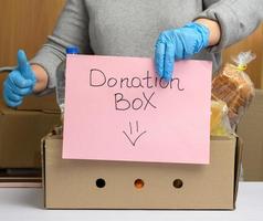 woman in gloves holds a sheet of paper with a lettering donation box and a cardboard box with food and things photo