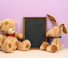 cute brown teddy bear and rabbit with long ears sit between empty wooden frame on purple background photo