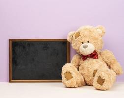 cute brown teddy bear sitting near empty wooden frame on purple background photo