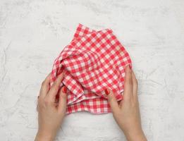 dos manos femeninas sostienen una servilleta de cocina textil a cuadros blanco-rojo arrugada sobre una mesa blanca foto