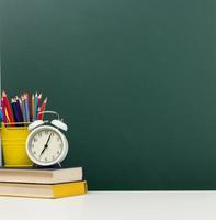 stack of books, a round alarm clock and multicolored pencils on the background of an empty green chalk board. Back to school photo