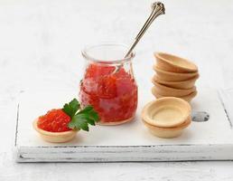 caviar chinook rojo en un frasco de vidrio y tartaletas redondas sobre una mesa blanca foto