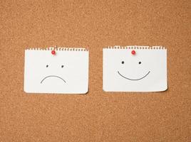 two paper leaves with a smile and a sad emotion attached with a button on a brown board photo