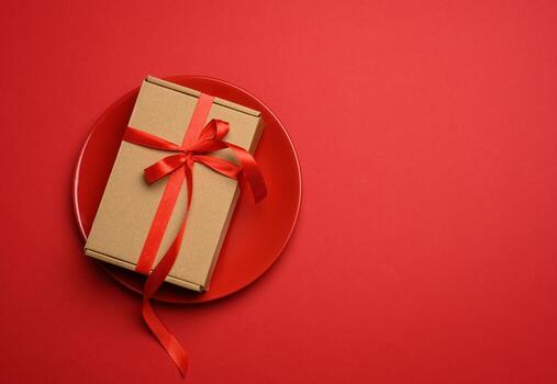 caja de cartón marrón rectangular atada con una cinta roja de seda yace en una placa roja de cerámica redonda foto