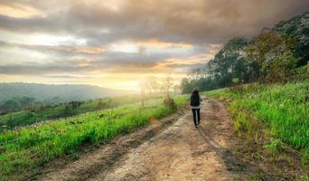 Rearview of relaxed woman walking on road in meadow field to travel in nature with morning sunlight sky. Rural scene. Outdoor activity in summer vacation. Landscape of green grass field and mountains. photo