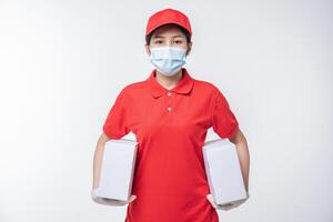 imagen de un joven repartidor consciente con gorra roja en blanco, camiseta uniforme, guantes de máscara facial de pie con una caja de cartón blanca vacía aislada en un estudio de fondo gris claro foto