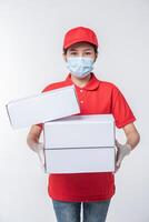 imagen de un joven repartidor consciente con gorra roja en blanco, camiseta uniforme, guantes de máscara facial de pie con una caja de cartón blanca vacía aislada en un estudio de fondo gris claro foto