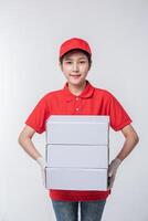 imagen de un joven repartidor con uniforme de camiseta en blanco con gorra roja de pie con una caja de cartón blanca vacía aislada en un estudio de fondo gris claro foto