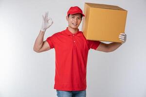 Image of a happy young delivery man in red cap blank t-shirt uniform standing with empty brown cardboard box isolated on light gray background studio photo