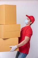 Image of a conscious young delivery man in red cap blank t-shirt uniform face mask gloves standing with empty brown cardboard box isolated on light gray background studio photo