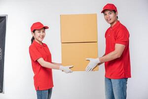 imagen de un joven y feliz repartidor con gorra roja, camiseta en blanco, uniforme de pie con una caja de cartón marrón vacía aislada en un estudio de fondo gris claro foto