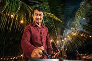 Portrait of Happy Asian young man cooking meat on barbecue grill at new year party. Bar-B-Q or BBQ on traditional stove. Night Party, people and celebration concept. photo