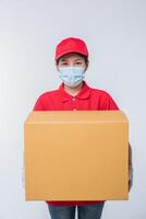 Image of a conscious young delivery man in red cap blank t-shirt uniform face mask gloves standing with empty brown cardboard box isolated on light gray background studio photo