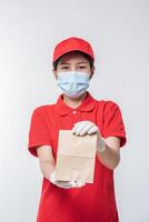 Image of a happy young delivery man in red cap blank t-shirt uniform face mask gloves standing with empty brown craft paper packet isolated on light gray background studio photo