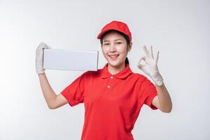 imagen de un joven repartidor con uniforme de camiseta en blanco con gorra roja de pie con una caja de cartón blanca vacía aislada en un estudio de fondo gris claro foto