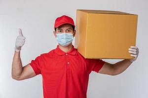 imagen de un joven repartidor consciente con gorra roja en blanco camiseta uniforme guantes de máscara facial de pie con una caja de cartón marrón vacía aislada en un estudio de fondo gris claro