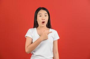 retrato de una joven asiática emocionada con una camiseta blanca señalando con el dedo a un lado aislada de fondo rojo foto
