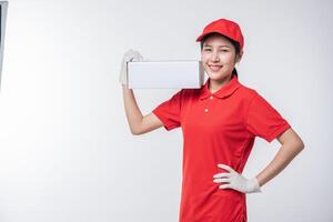 imagen de un joven repartidor con uniforme de camiseta en blanco con gorra roja de pie con una caja de cartón blanca vacía aislada en un estudio de fondo gris claro foto