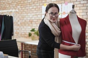 Asian mature female fashion designer stands near dressing puppet, looking at camera with smile in studio, proudly works with clothing design collection, professional boutique tailor SME entrepreneur. photo