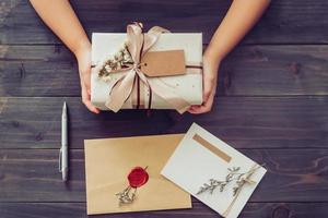 Above of hand girl present craft gift box with card and pen on wooden table background photo