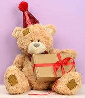 lindo oso de peluche marrón con una gorra roja se sienta y sostiene una caja marrón con un regalo foto