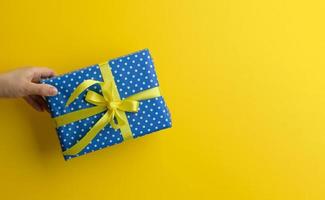 mano femenina sostiene una caja de regalo azul sobre un fondo amarillo, concepto de feliz cumpleaños foto