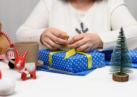 woman in a white sweater sits at the table and wraps gifts for Christmas. Preparing for the holidays photo