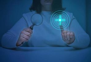 a woman sits at a table and holds a magnifying glass, with the other hand presses on a virtual screen with neon circles. Search for information, start the program photo