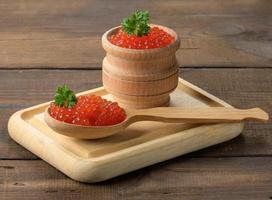 el caviar rojo de salmón rosado se encuentra en una cuchara de madera sobre una tabla para cortar. mesa de madera marrón foto