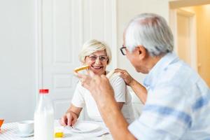 Cheerful senior couple having breakfast at home. Elderly man hugging woman. Mature woman holding piece of bread. An old man and woman sitting at the table, talking. photo