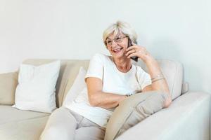 hermosa anciana caucásica conversando por teléfono con su vieja amiga usando un dispositivo electrónico, discutiendo las últimas noticias con una linda sonrisa en la cara, sentada en casa foto