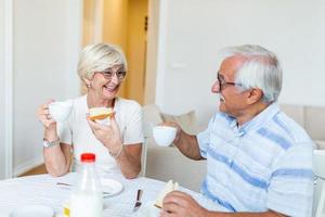 Senior couple having breakfast and drinking coffee. Elderly couple having their meal at home. Mature woman holding piece of bread. An old man and woman sitting at the table, relaxing. photo