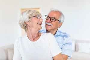 Elderly couple standing in the living room. Portrait of happy senior couple embracing each other in living room at home. Old couple looking each other with love and smilling. photo