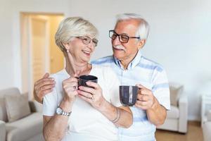 Happy senior couple standing in the living room. Looking each other smilling and holding cup of coffee or tea. Elderly husband hugging his lovely wife photo